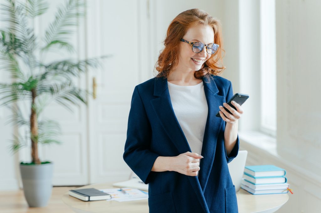 Positive red haired woman checks e-mail box on modern smartphone device